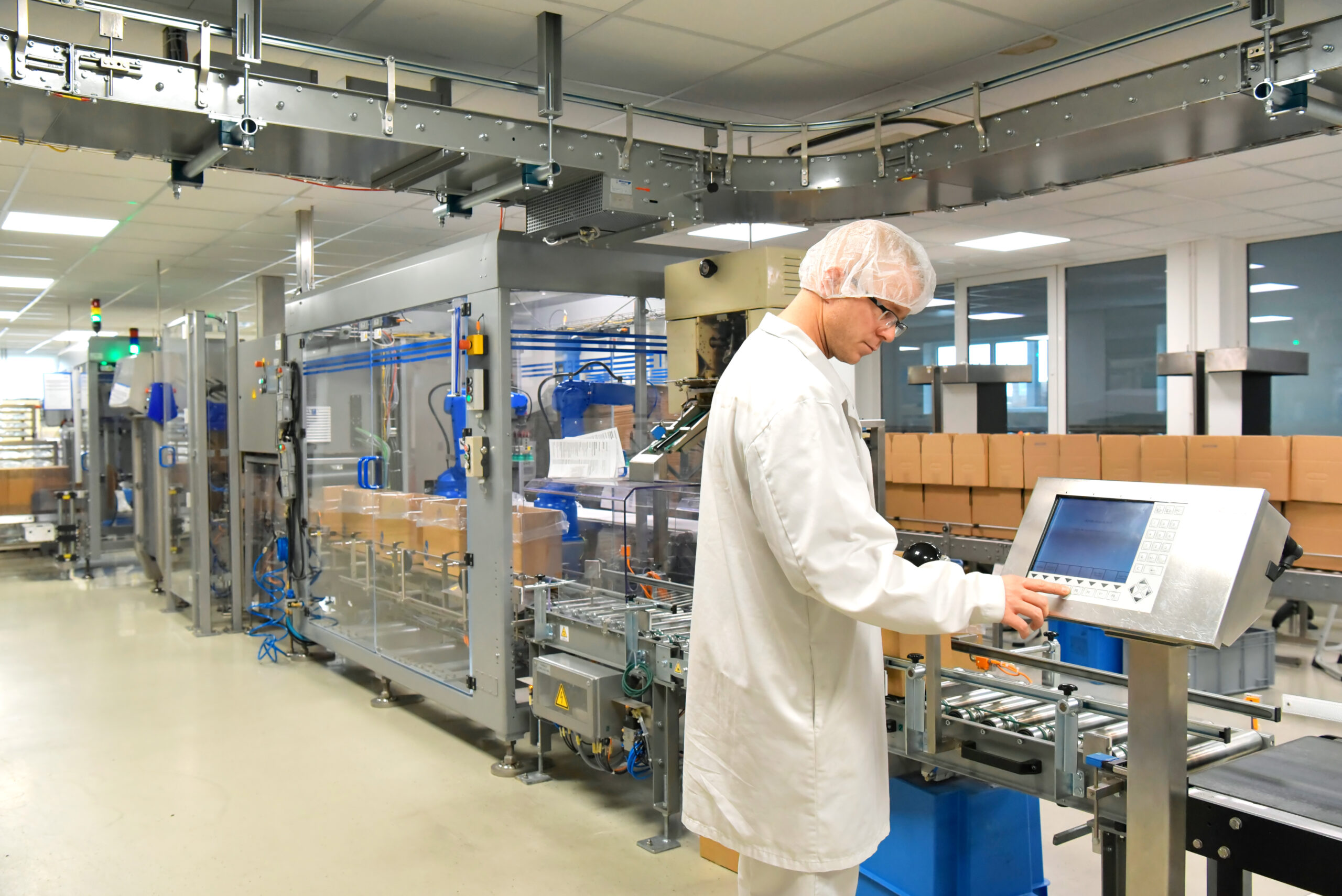 Person wearing a white coat and hair net using the control panel for an automated conveyor with boxes on it